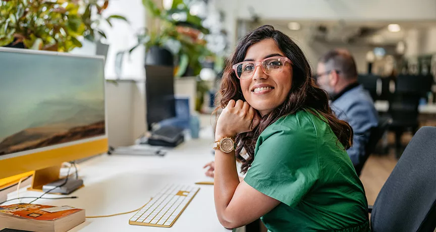 mulher sorrindo para a câmera enquanto trabalha em frente a computador.
