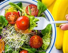 Imagem mostra uma salada com tomates e frutas ao fundo