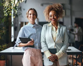 duas mulheres em ambiente corporativo segurando cadernos sorrindo enquanto olham para a câmera.