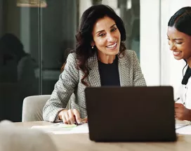 duas mulheres sorrindo enquanto conversam em frente a um notebook no escritório.