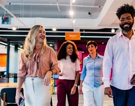 time de três mulheres e um homem caminhando, conversando e rindo em escritório.