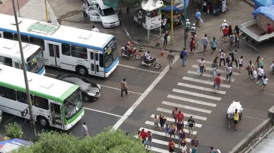 Foto mostra o cruzamento de uma avenida movimentada de cima, com a faixa de pedestre e ônibus e carros esperando no semáforo