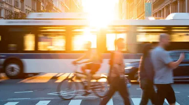Foto mostra um cruzamento de avenida, com pedestres passando na faixa e um ônibus de fundo