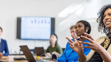 mulher explicando algo para uma sala de reunião com várias pessoas e apresentação ao fundo.