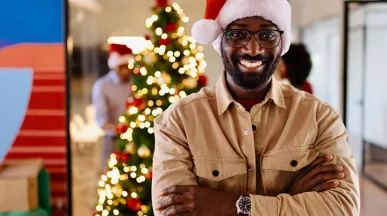 homem com chapéu de papai noel sorrindo na empresa com árvore de natal ao fundo.