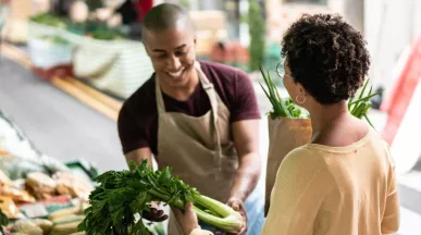A imagem mostra uma mulher comprando legumes na feira, com um vendedor atendendo ela