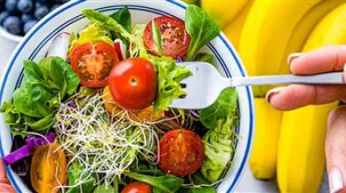 Imagem mostra uma salada com tomates e frutas ao fundo