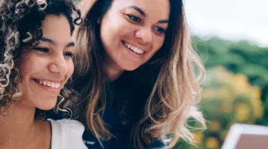 duas mulheres sorrindo enquanto olham algo em computador ao ar livre.