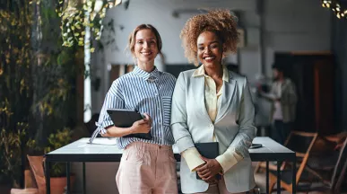 duas mulheres em ambiente corporativo segurando cadernos sorrindo enquanto olham para a câmera.