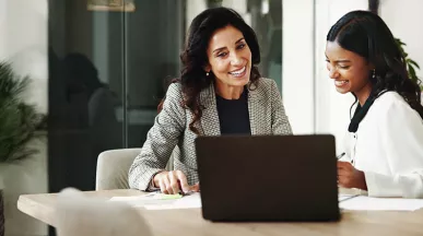 duas mulheres sorrindo enquanto conversam em frente a um notebook no escritório.