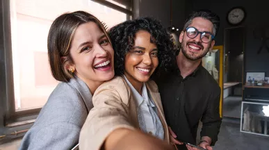 Foto mostra três colegas de trabalho tirando uma selfie no escritório