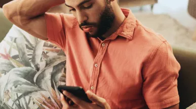 Pessoa sentada em um sofá, usando uma camisa de cor salmão, olhando para um smartphone que segura com as duas mãos. Ao lado há almofadas decorativas, incluindo uma com estampa de folhas. O ambiente interno é aconchegante, com iluminação suave e móveis desfocados ao fundo.