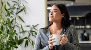A imagem mostra uma mulher, em casa, segurando uma caneca e sorrindo olhando para a janela