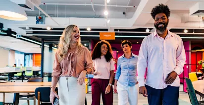 time de três mulheres e um homem caminhando, conversando e rindo em escritório.