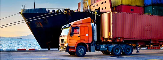 Foto mostra um grande caminhão saindo do porto, com um navio de carga ao fundo