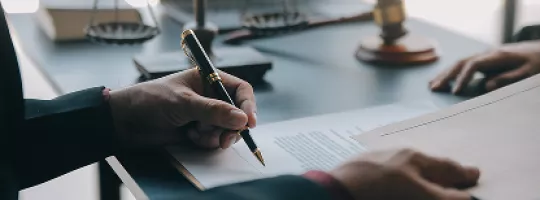 Pessoa assinando um documento em mesa com itens jurídicos ao fundo.