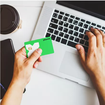 Pessoa usando laptop e segurando cartão Pluxee, com celular e café sobre a mesa
