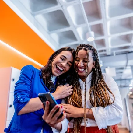 Foto mostra duas colegas de trabalho vendo o smartphone