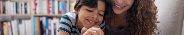 Pessoa adulta e criança colocando moedas em cofrinho em casa, com livros ao fundo.